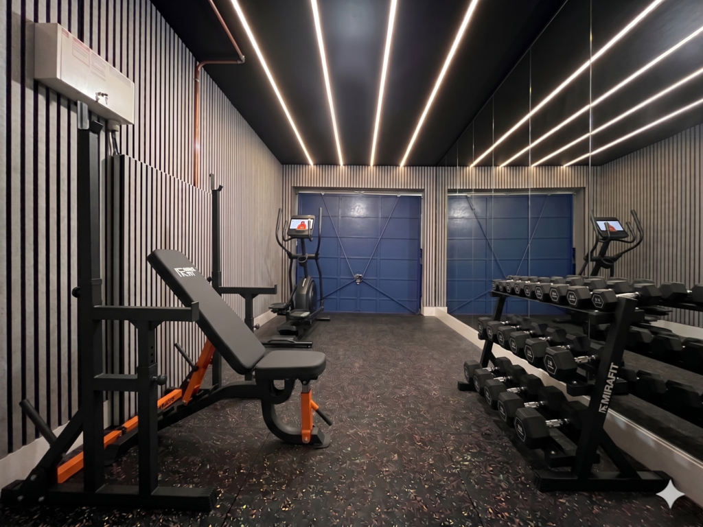 Finished UK garage home gym featuring an adjustable bench and squat rack, hex dumbbell rack, elliptical cross trainer, mirrored wall and warm strip lighting overhead