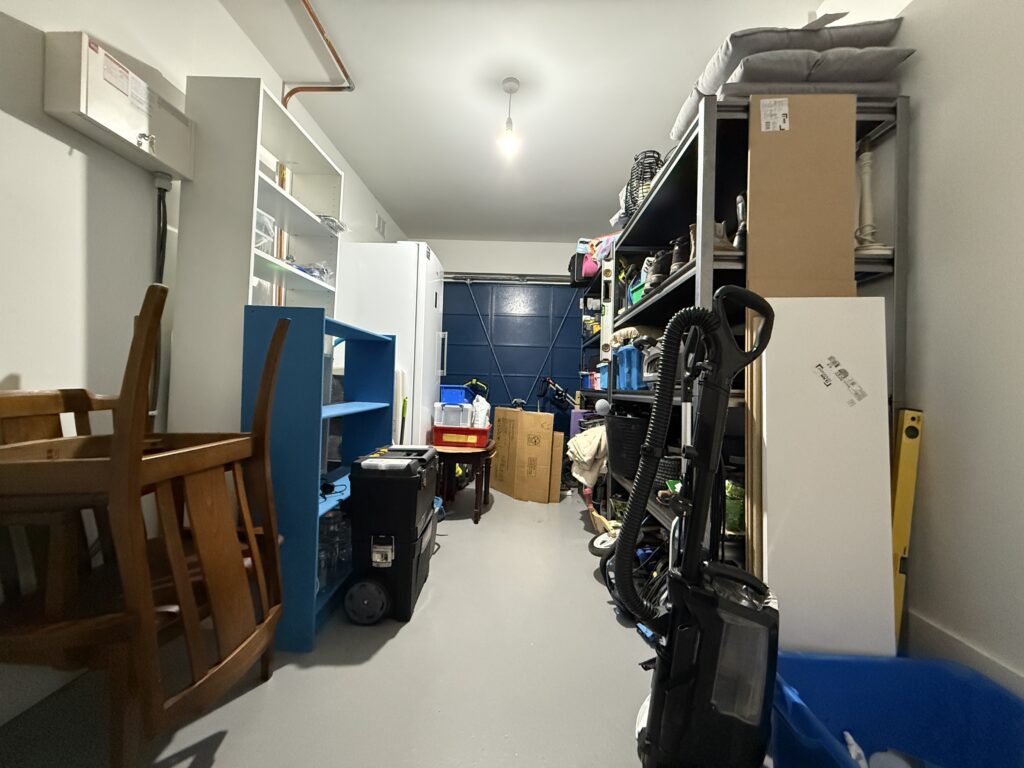 Cluttered single garage in the UK filled with shelving, storage boxes, a vacuum cleaner, chairs and general household overflow before the home gym conversion begins