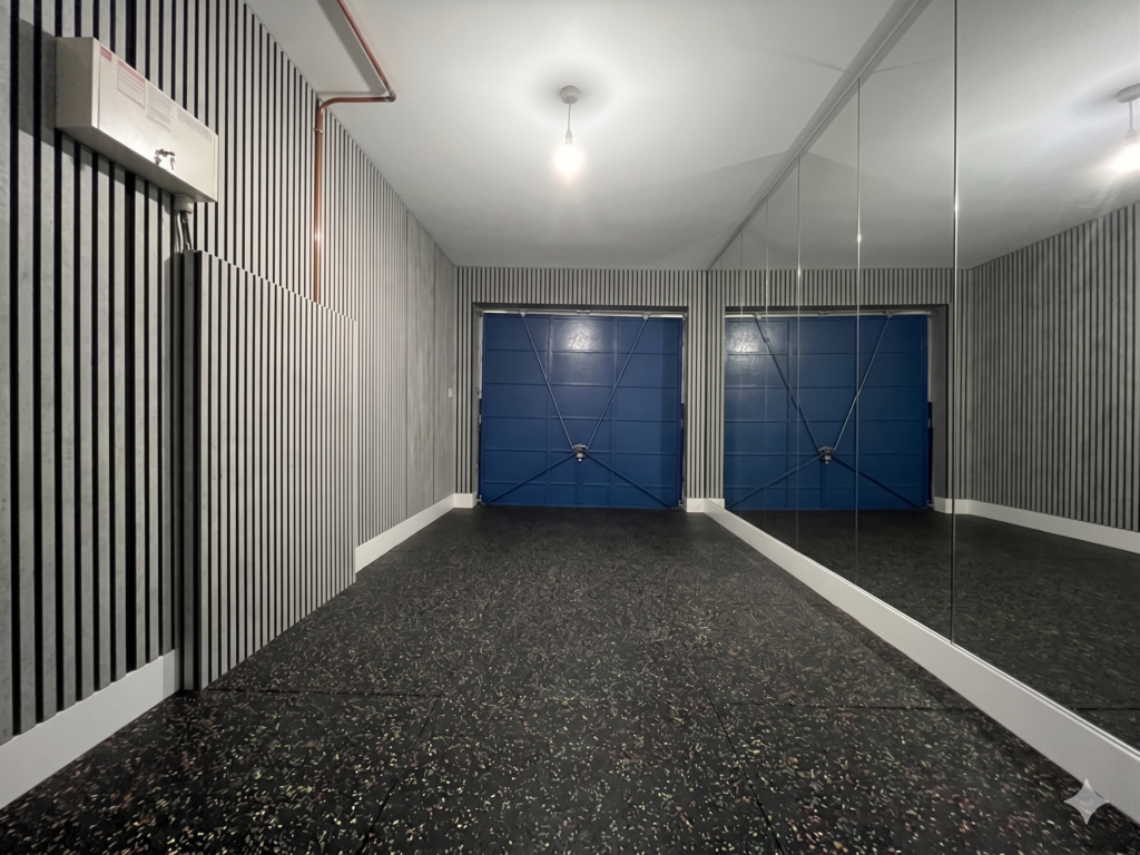 Full length mirrored wall reflecting grey wood acoustic panels and black rubber flooring inside a small UK garage being converted into a home gym