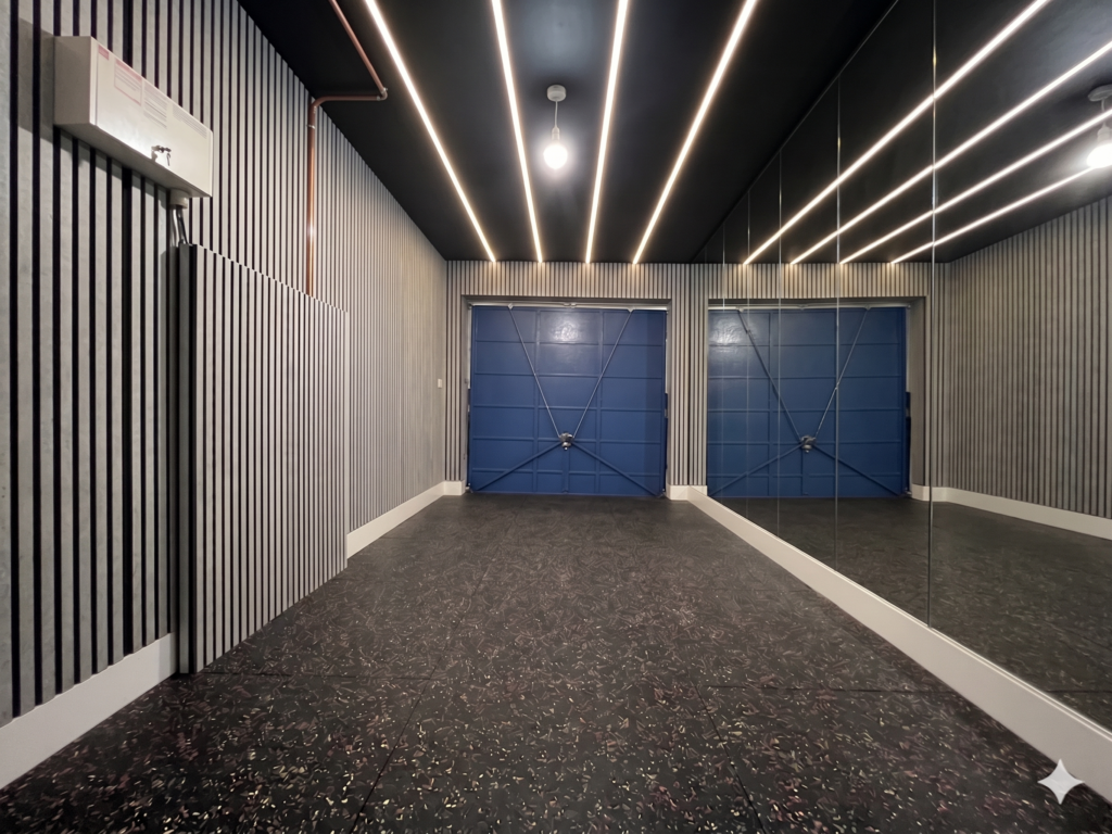 Small UK garage home gym with a black painted ceiling, fanning warm LED strip lights, mirrored wall, grey acoustic wood panelling and black rubber flooring