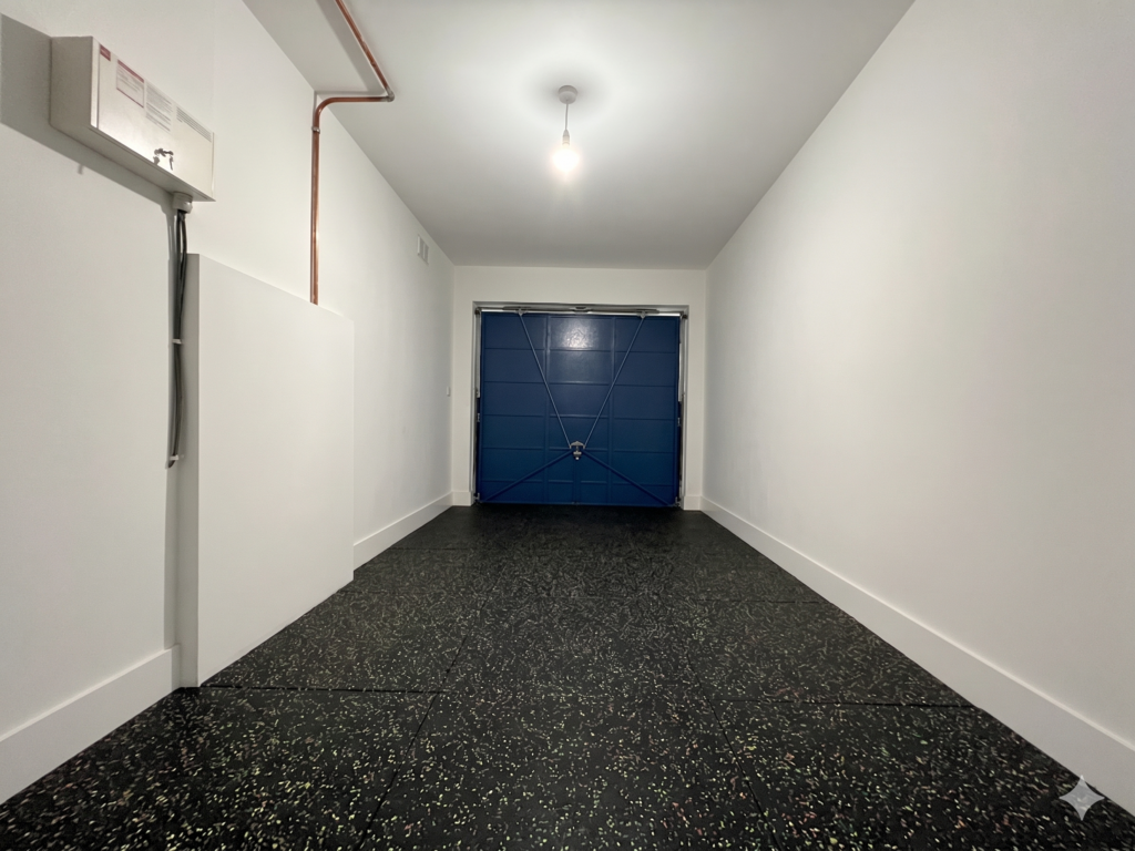 Black speckled rubber gym flooring laid across the full length of a narrow UK garage with white walls and a blue garage door at the far end