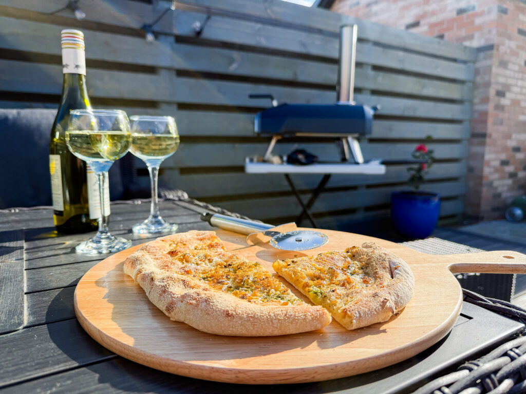 Two large slices of garlic bread pizza on a wooden board with wine glasses and the Ooni Karu 2 oven setup in the background of a modern garden.