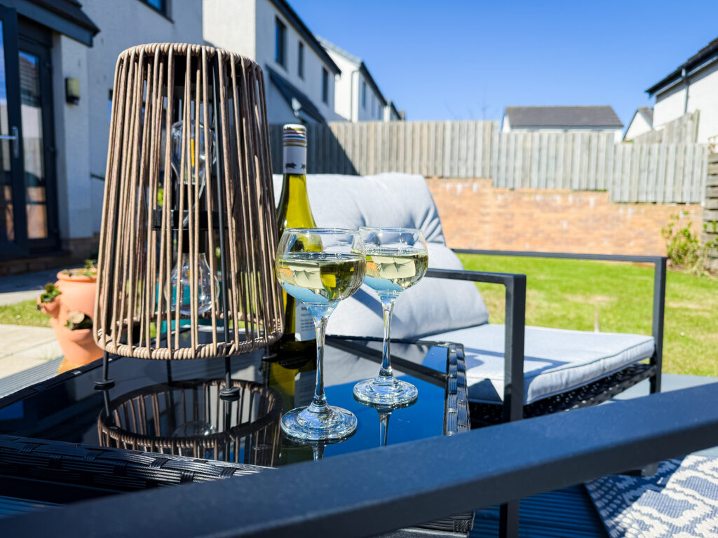 Two glasses of white wine and a bottle on a black glass-top garden table under a clear blue sky.