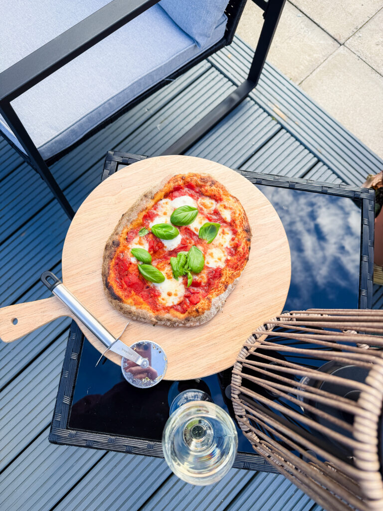 An overhead shot of a whole margherita pizza on a wooden board, surrounded by a pizza cutter, white wine, and outdoor garden furniture.