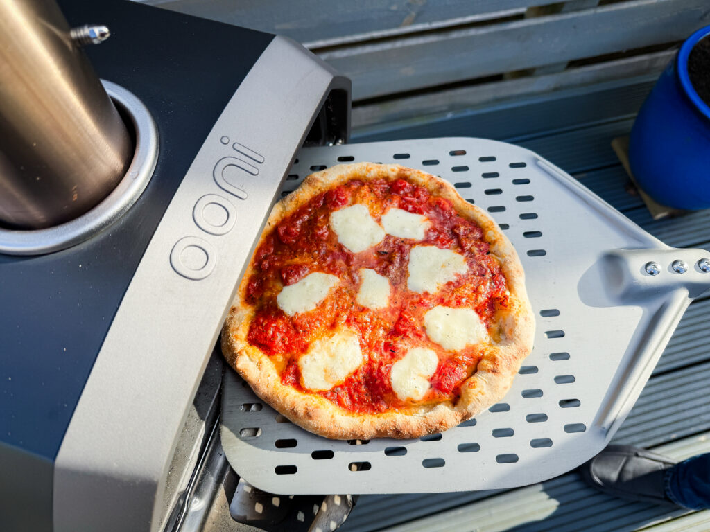 An overhead perspective of a pizza being launched into the mouth of an Ooni Karu 2 oven using a metal perforated peel.