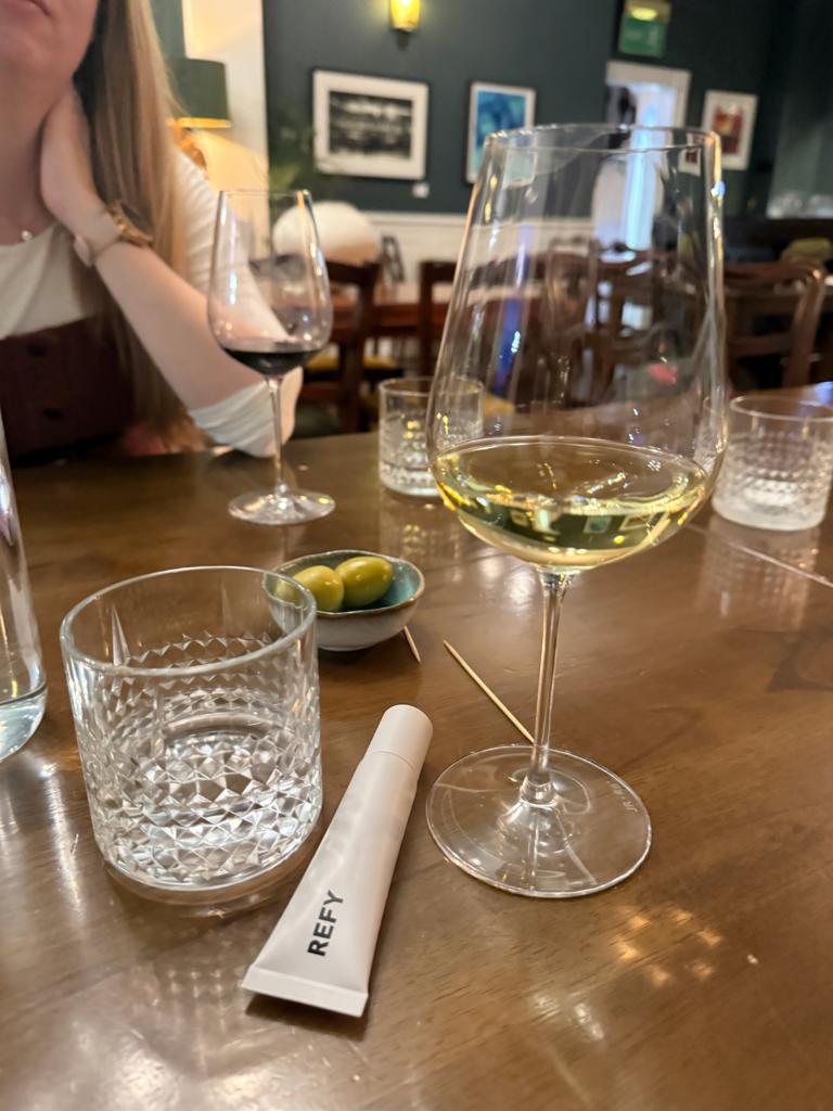A minimalist white tube of Refy Lip Jelly placed on a restaurant table next to a glass of white wine and a small bowl of green olives.