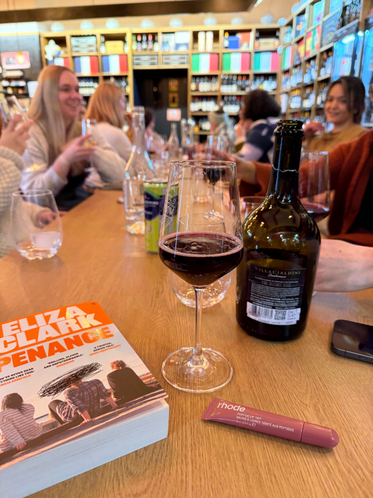 A tube of Rhode Peptide Lip Tint in a mauve shade sitting on a wooden table next to a glass of red wine and a copy of the book Penance by Eliza Clark.