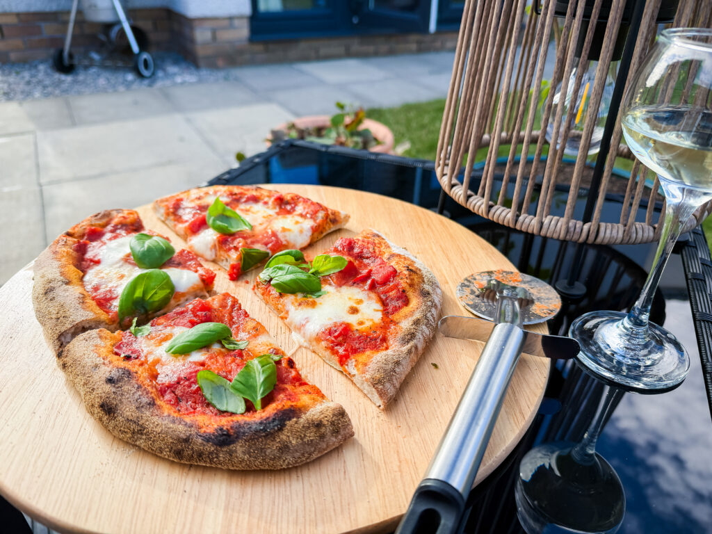 Slices of margherita pizza with fresh basil on a wooden board next to a pizza cutter and a glass of white wine on a glass garden table.