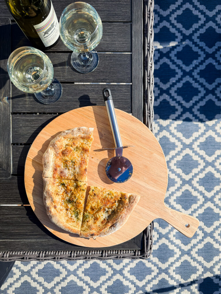 A top-down shot of garlic bread pizza on a wooden board, a pizza cutter, and two glasses of chilled white wine on a dark slatted garden table.