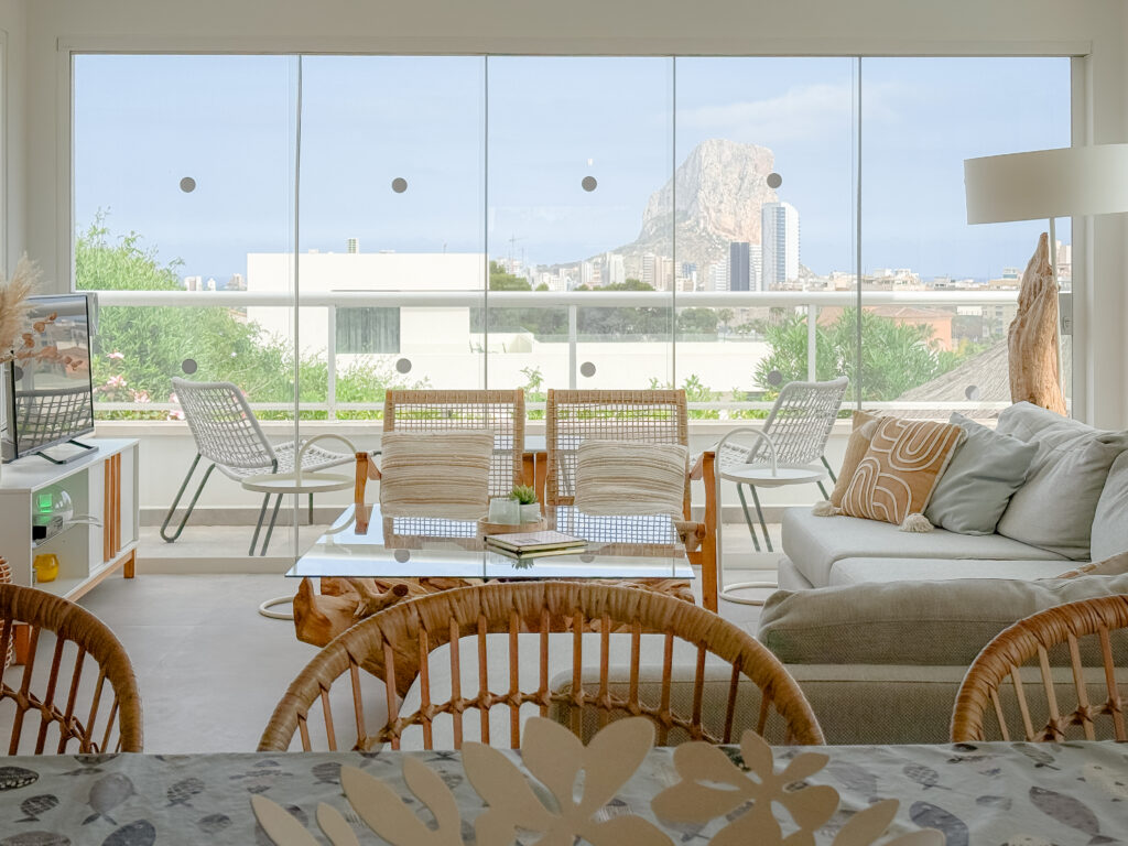 A stunning framed view of the Peñón de Ifach rock formation through large glass living room windows, with modern rattan furniture in the foreground.