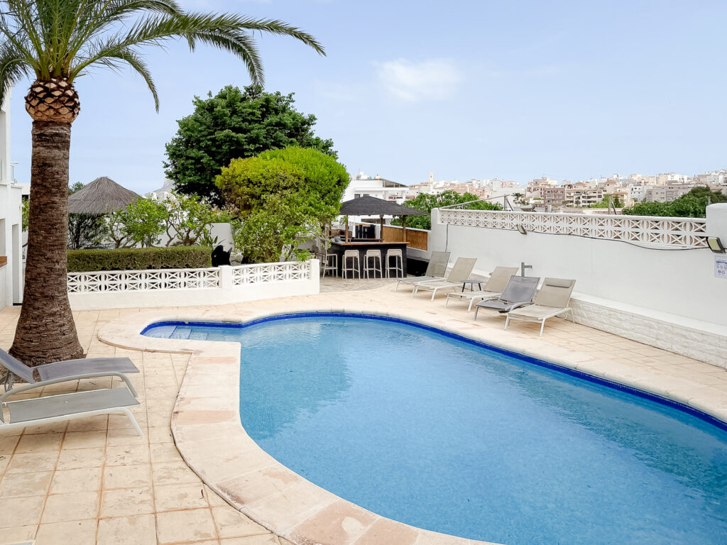 A bright, kidney-shaped swimming pool in a Spanish villa with sun loungers, a palm tree, and a view of the white buildings of Calpe in the distance.