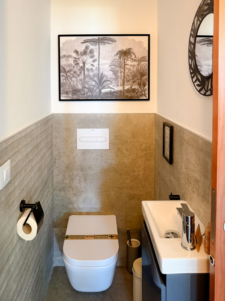 A narrow, modern powder room with grey textured wall tiles, a wall-mounted toilet, a small sink, and tropical-themed wall art.