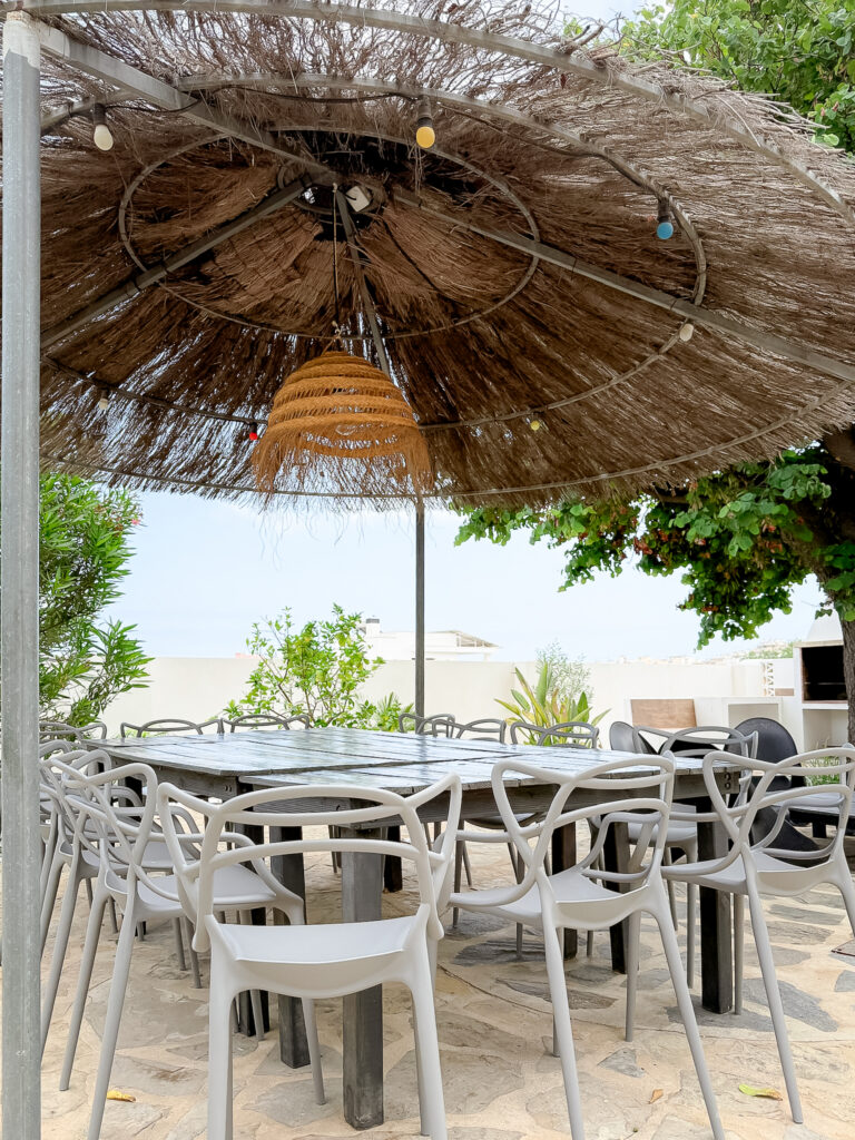 A long black wooden dining table with grey designer chairs, positioned under a large tropical-style thatched umbrella with a view of the sea.