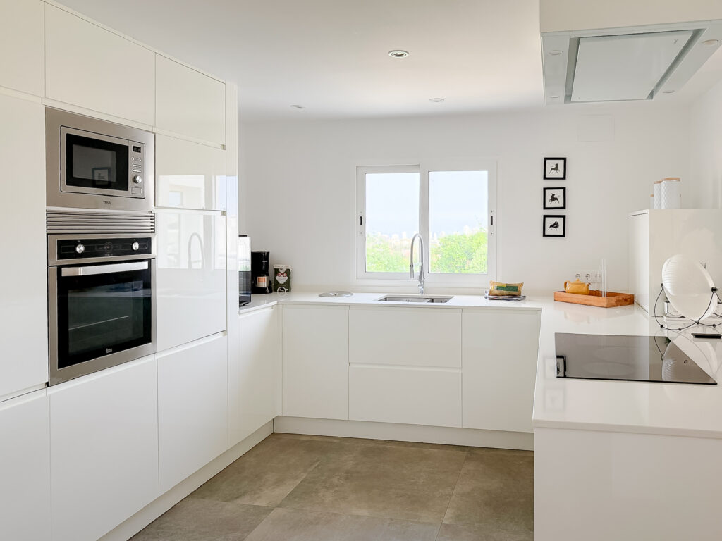 A clean, wide-angle view of a white modern kitchen with an island, stone-effect floor tiles, and a large window overlooking greenery.
