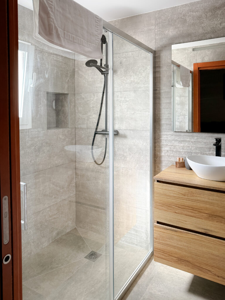 A sleek, modern bathroom featuring large grey stone-effect tiles, a glass walk-in shower, and a light wood vanity with a white vessel sink.
