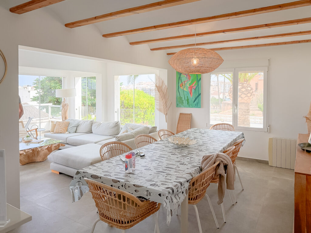 An open-plan indoor living and dining room with wooden ceiling beams, a long dining table, and large glass doors opening to a coastal view.