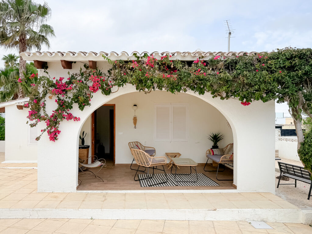 A white arched outdoor sitting area draped in bright pink bougainvillea flowers, with rattan chairs and a striped rug.