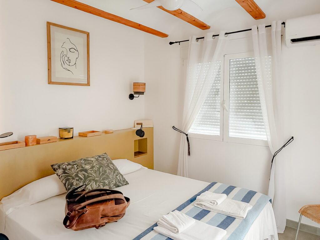 A bright, Mediterranean-style bedroom with white walls, wooden ceiling beams, white linens, and a tan headboard.