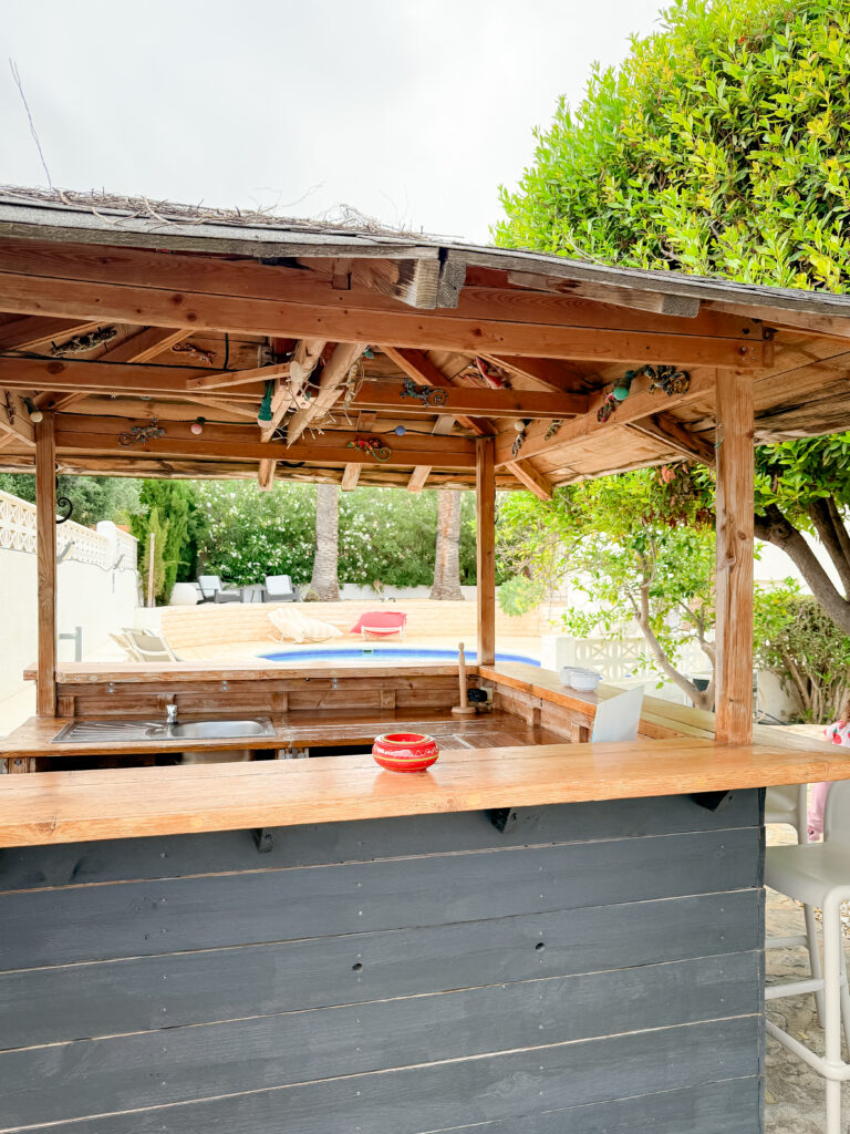 A rustic, thatched-roof outdoor pool bar with wooden countertops and a small sink, looking out toward the swimming pool and greenery.