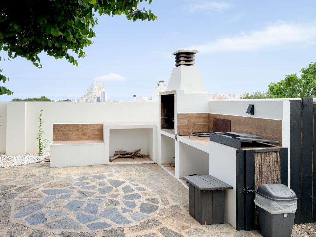 A purpose-built, white-rendered outdoor cooking station with a wood-fired grill, chimney, and stone patio, overlooking the Peñón de Ifach.
