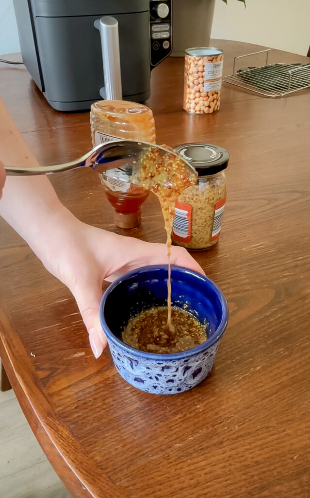 A spoonful of textured wholegrain mustard being lifted over a blue patterned ceramic bowl, with jars of honey and mustard on the table behind it.