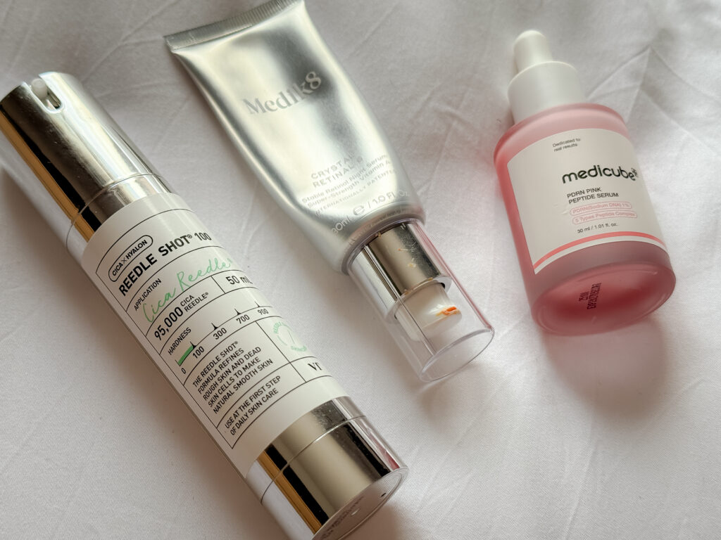 Three top trending skincare active products lying on a white sheet: VT Cosmetics Reedle Shot 100, Medik8 Crystal Retinal 6 tube, and Medicube PDRN Pink Peptide Serum bottle.