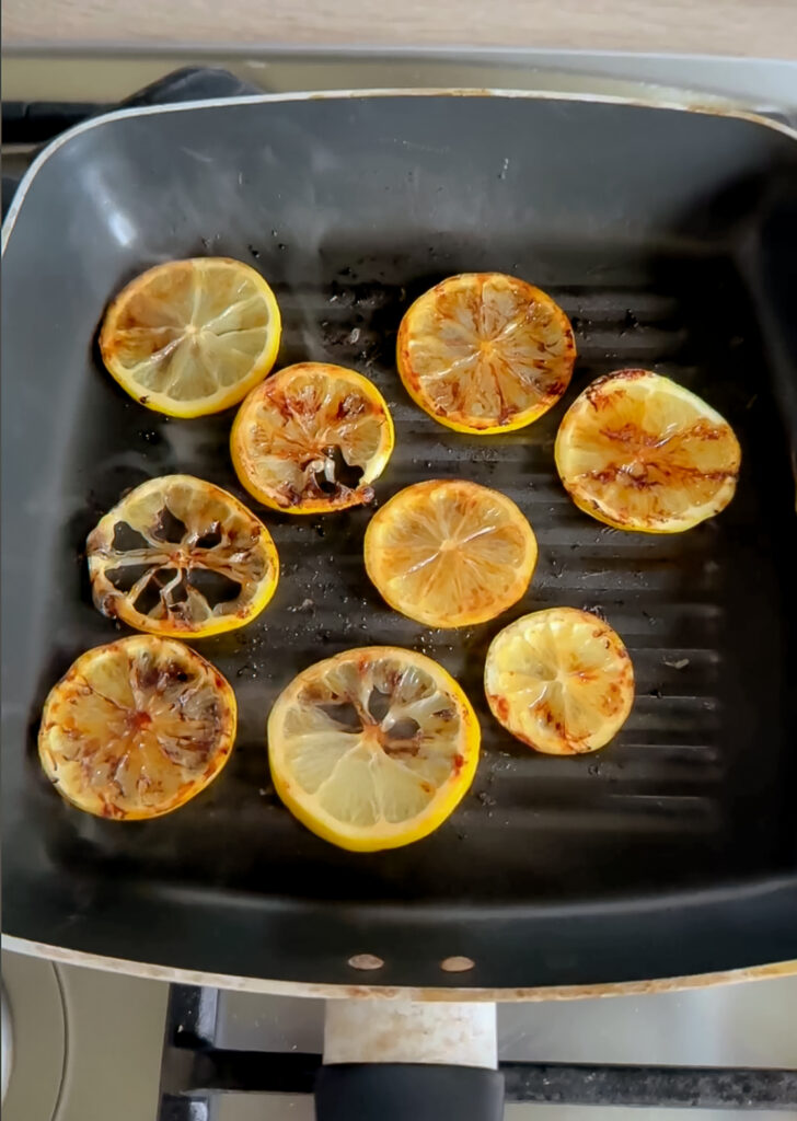 Slices of fresh lemon searing and charring face-down in a hot, dark ridged griddle pan on the stovetop.