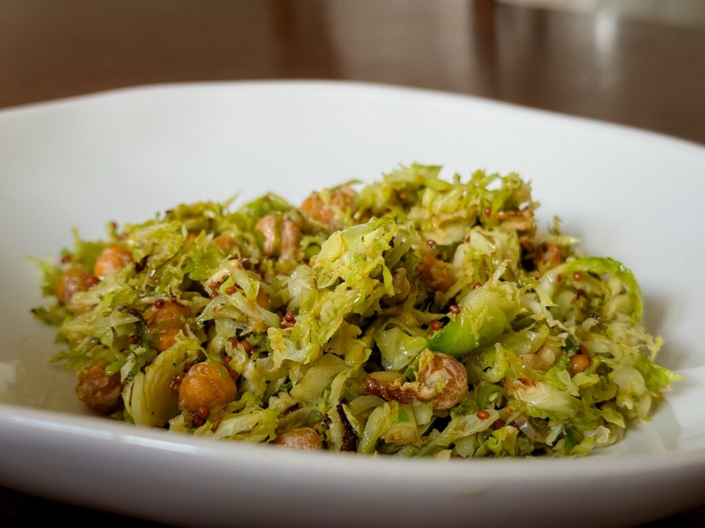 A close-up shot of the finished Brussels sprout salad served in a wide white ceramic bowl. The salad features finely shredded, golden-edged air-fried Brussels sprouts tossed with roasted chickpeas and coated in a textured wholegrain mustard dressing.
