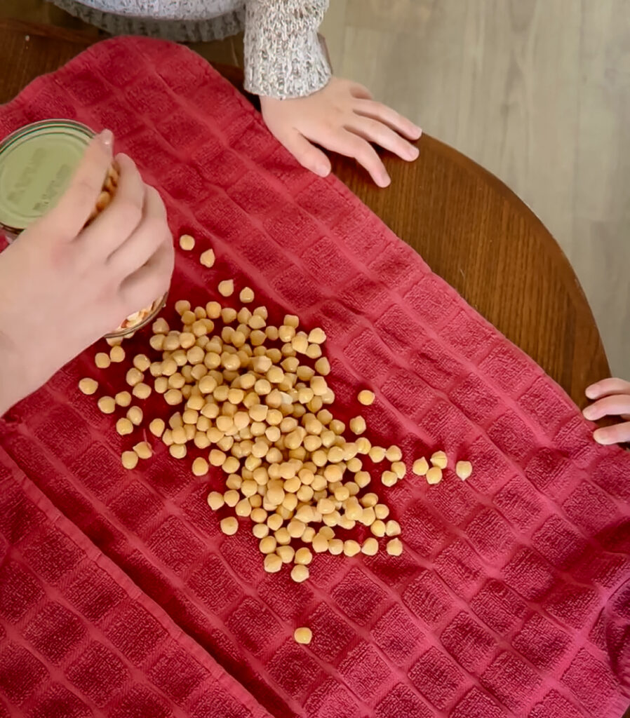 Canned chickpeas being poured out onto a red textured kitchen towel to dry, with a child's hands resting on the wooden table nearby.