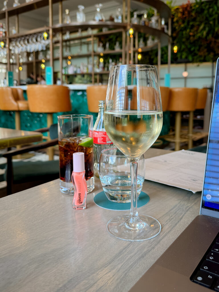 A single pink tube of Gisou Honey Infused Lip Oil stands upright on a restaurant table next to a glass of white wine and a laptop, showcasing its signature wavy bottle design.