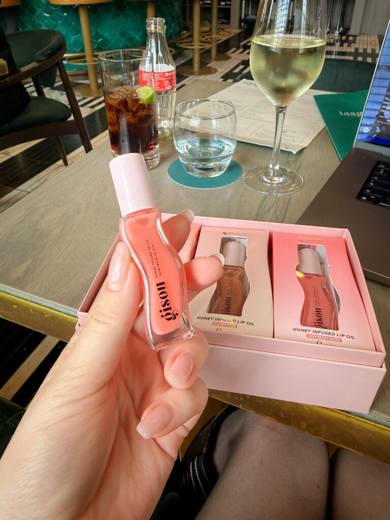 A close-up of a hand with a manicured neutral nail holding a pink Gisou lip oil bottle over an open pink gift set containing shades Glazed Plum and Raspberry Swirl.