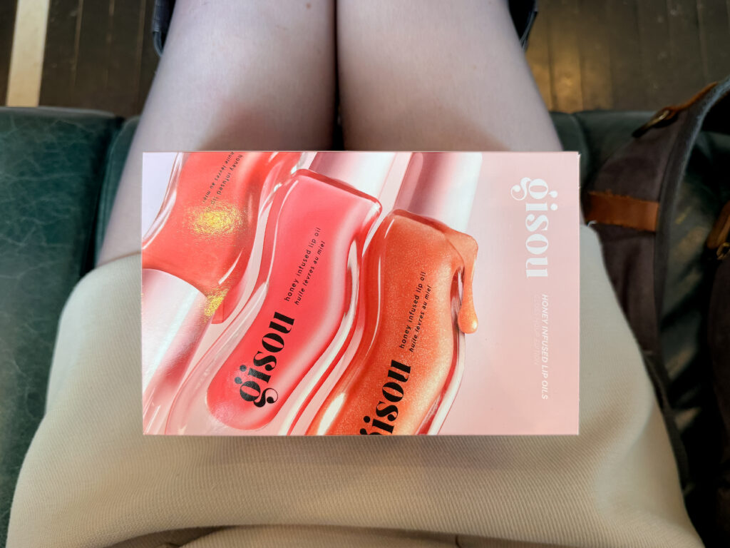 Top-down view of the Gisou Honey Infused Lip Oils Gloss House Edition gift box resting on a person's lap, featuring vibrant swatches of red, pink, and shimmering gold lip oils printed on the cover.