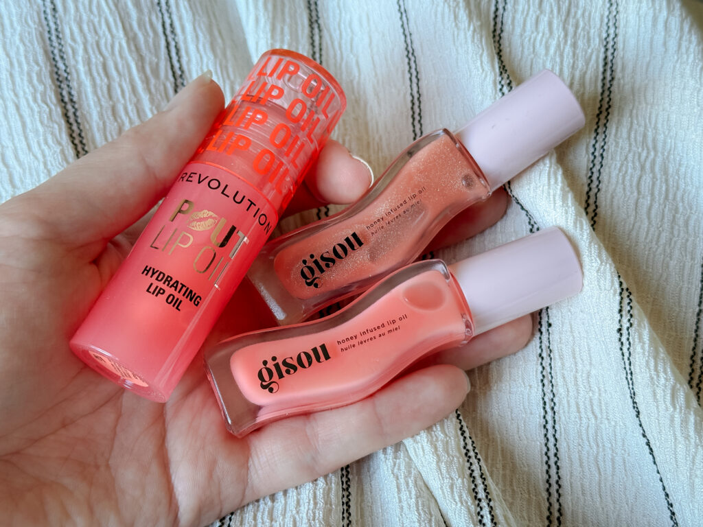 A hand holding three pink lip oil products against a striped fabric background, including one Revolution Pout Lip Oil tube and two shades of Gisou Honey Infused Lip Oil.