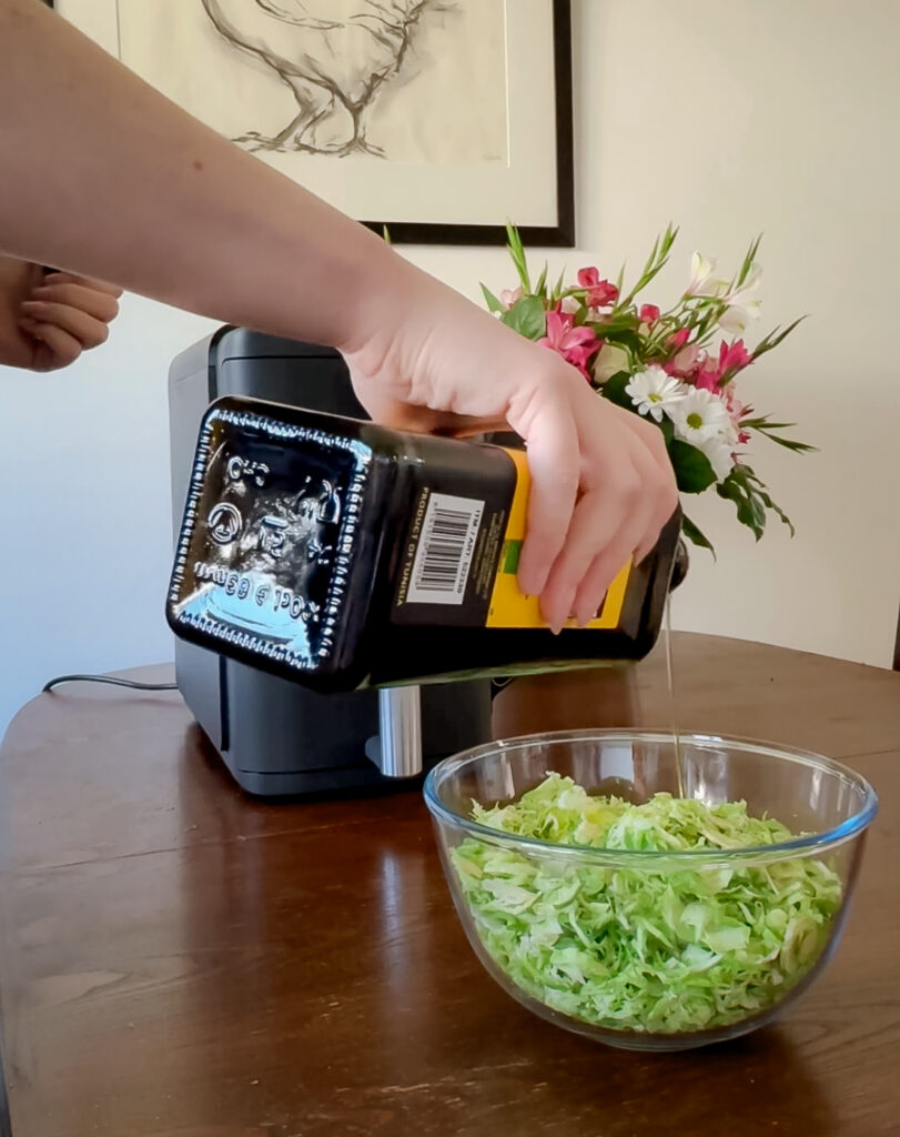 A person pouring olive oil from a dark glass bottle into a clear glass bowl filled with finely shredded raw Brussels sprouts.