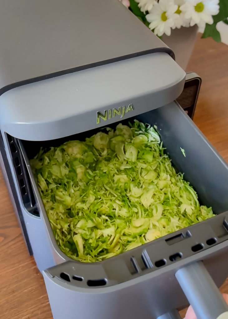 A close-up view looking into an open grey Ninja air fryer drawer packed with seasoned, shredded raw green Brussels sprouts.