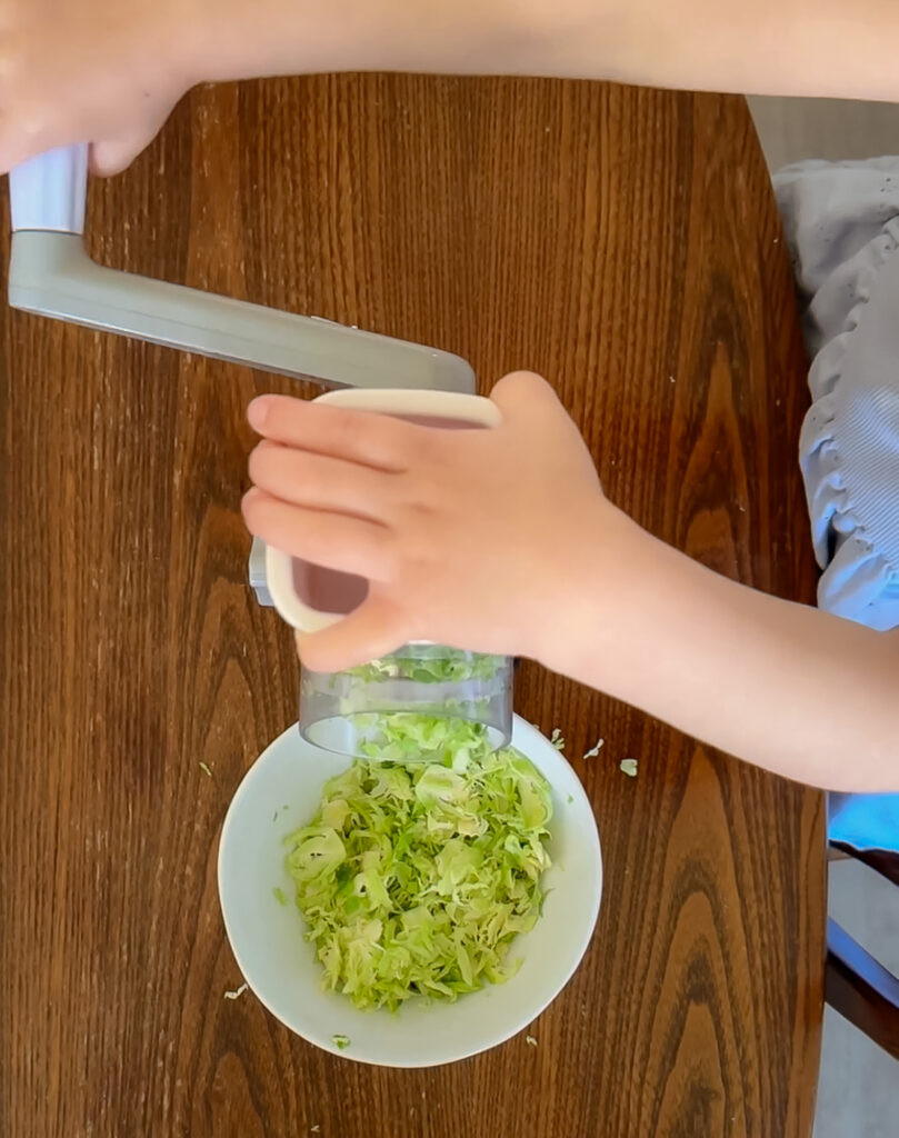 An overhead view of a child using a grey rotary mandoline slicer to finely shred green Brussels sprouts into a white ceramic bowl on a wooden table.