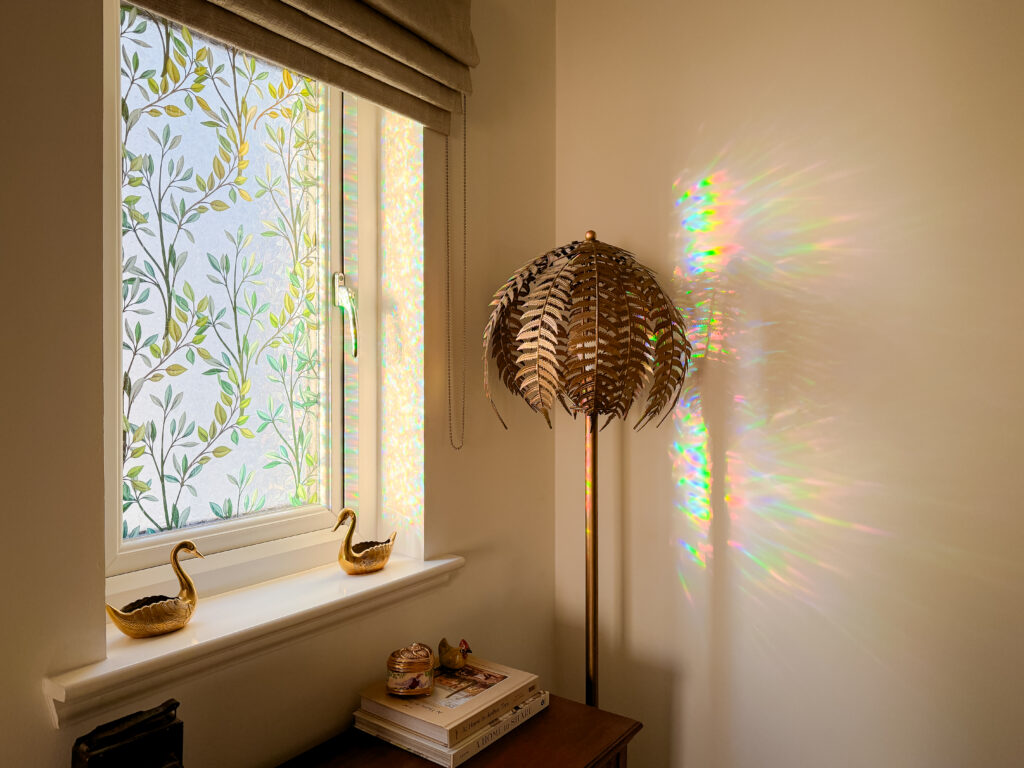 A hallway window fitted with a botanical leaf pattern stained glass window film. Sunlight filters through the decorative privacy vinyl, casting a vibrant, shimmery rainbow light reflection across the adjacent beige wall next to a gold brass palm tree floor lamp.