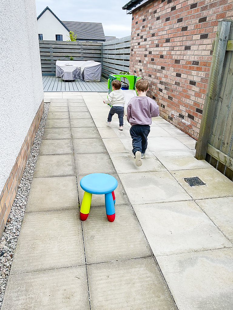 Two children running away across a newly levelled grey slabbed patio in a new build garden.
