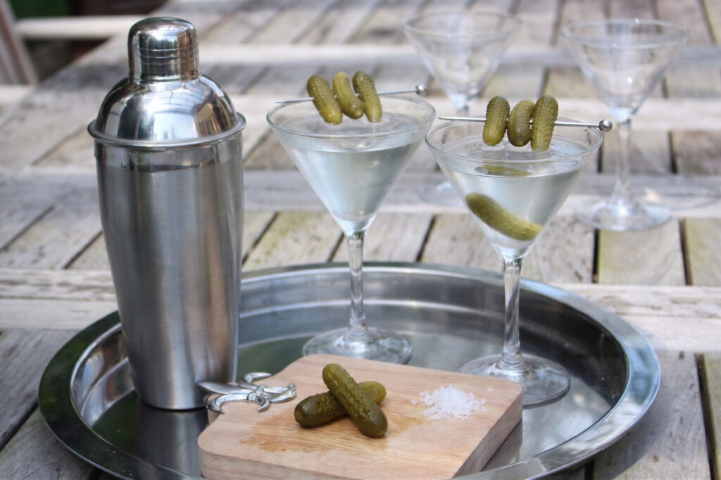 pickle martini next to a shaker on an outdoor table in the sun with pickle garnish
