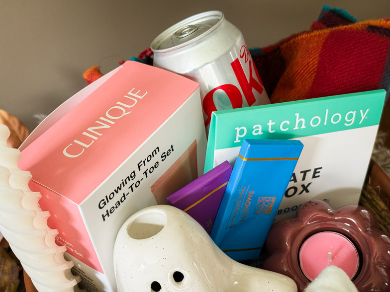 A close up of a filled boo basket showing a Clinique gift set, Patchology masks, chocolate bars and a tartan blanket