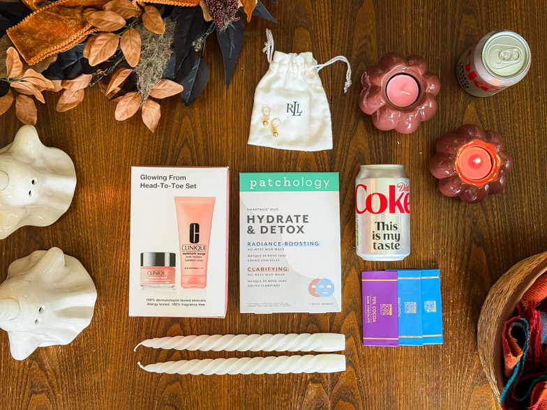 Boo basket contents laid out on a wooden table including Clinique skincare, Patchology mud masks, gold earrings, chocolate and pumpkin candle holders