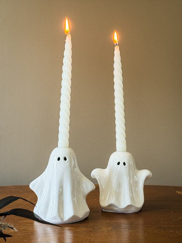 Two white ceramic ghost candle holders with lit white twisted taper candles against a warm neutral wall